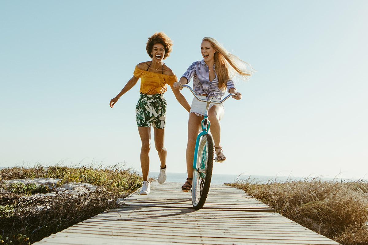 Excited woman riding bike down the boardwalk with her friends running along. Two female friends having a great time on their vacation. Panoramic shot with lots of copy space on background.