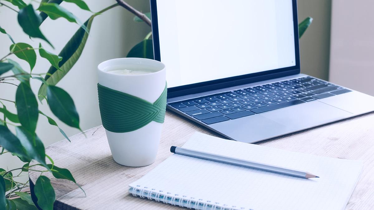 A modern office workplace surrounded by green plants. A wooden table with a laptop, notebook, pencil and a cup of water and lemon. Desk plants and flowers have many benefits, according to the latest research.