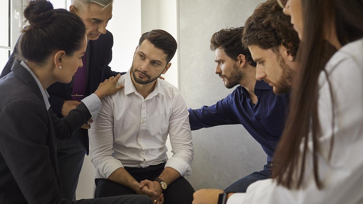 Group of different people sitting in a circle supporting and comforting a sad upset young man. Concept of psychological help, group therapy and overcoming problems together