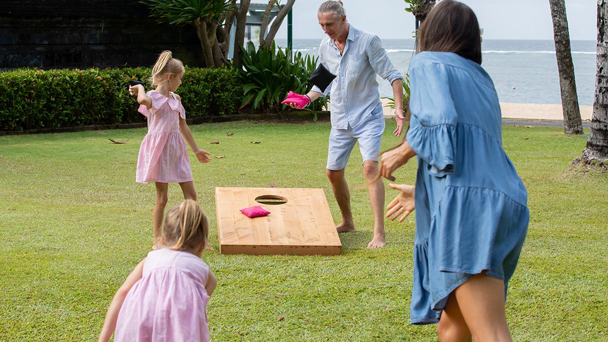Happy family playing cornhole game by the sea on sunny summer day. Parents and children playing bean bag toss