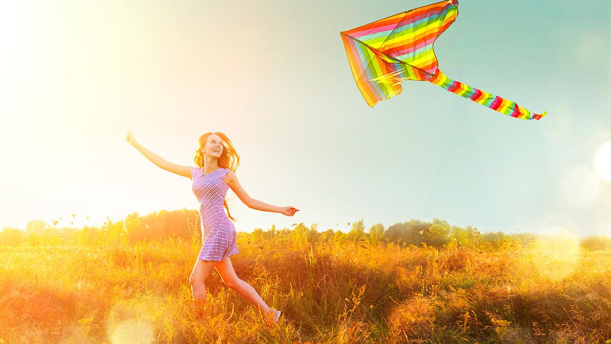 Beauty girl in short dress running with flying colorful kite ove