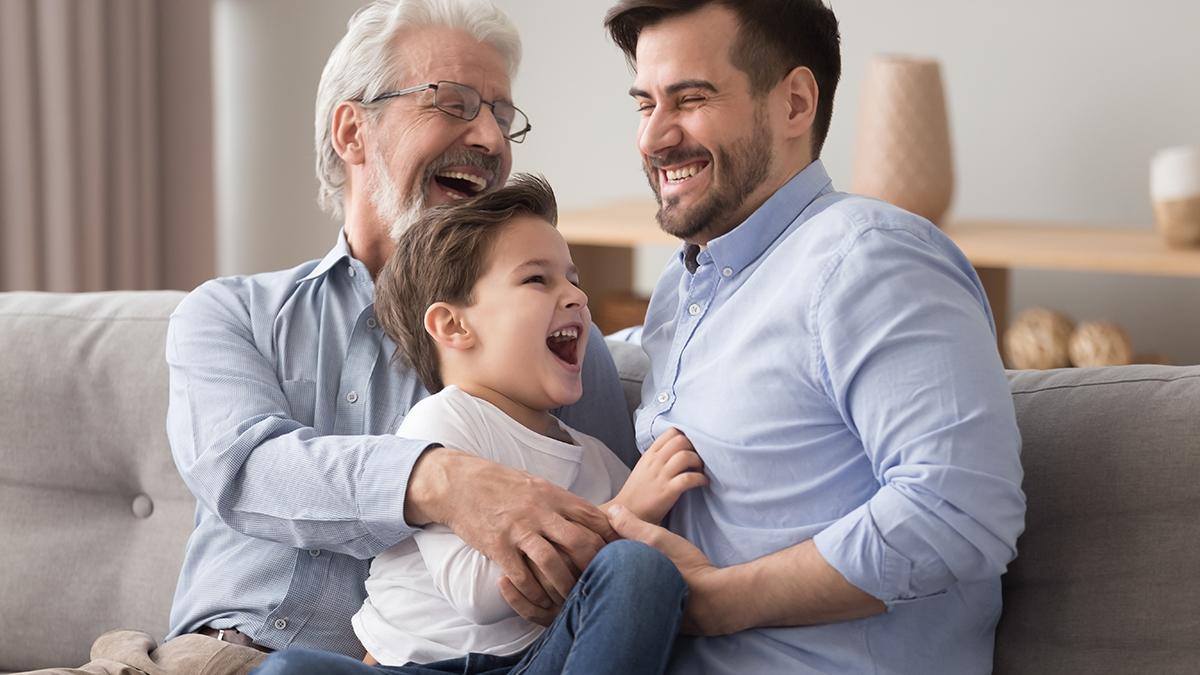 Happy grandfather and father tickling little boy, three generations of men having fun together, sitting on couch, laughing grandparent with son and grandchild relaxing spending weekend at home